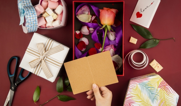 top view of female hand with a small open postcard over the gift box with coral color rose flower with scattered petals and a box filled with marshmallow on red background